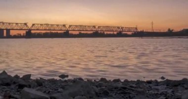 Şehir nehir kıyısının 4K UHD zaman çizelgesi. Güneş ışınları, mavi gökyüzü ve ufukta demiryolu köprüsü. Düz küçük dalgalar, su ve motorlu kaydırma hareketleriyle yaz gündoğumu zamanı
