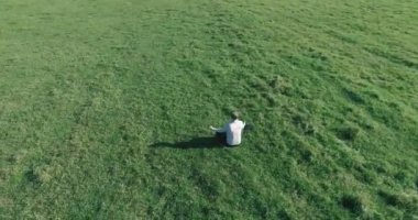 Havadan bakış açısı. Yeşil çimlerde oturan ve kırsal alanda not defteri olan bir adamın etrafında alçak yörünge uçuşları. Günlük giysi ve zen meditasyon pozu. Güneş ışınları. Radikal hareket.