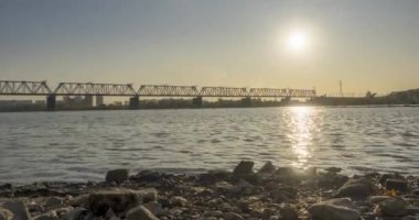 Şehir nehir kıyısının 4K UHD zaman çizelgesi. Güneş ışınları, mavi gökyüzü ve ufukta demiryolu köprüsü. Düz küçük dalgalar, su ve motorlu kaydırak hareketleriyle yaz günbatımı zamanı