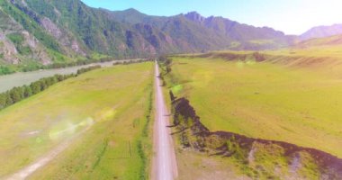 Hava UHD 4K: güneşli yaz sabahında kırsal dağ yolu ve çayır üzerinde havadan uçuş. Ufukta yeşil ağaçlar ve güneş ışınları var. Özgür kırsal asfalt karayolu ve nehir.