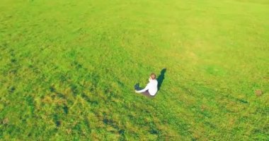 Havadan bakış açısı. Yeşil çimlerde oturan ve kırsal alanda not defteri olan bir adamın etrafında alçak yörünge uçuşları. Günlük giysi ve zen meditasyon pozu. Güneş ışınları. Radikal hareket.