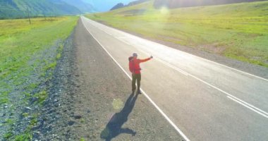 Hava 4K UHD görüntüsü. Asfalt yolda yürüyen otostopçu turist üzerinde alçak irtifa uçuşu. Büyük bir kırsal vadi ve yaz günü güneşli bir çayır. Sırt çantalı gezgin..