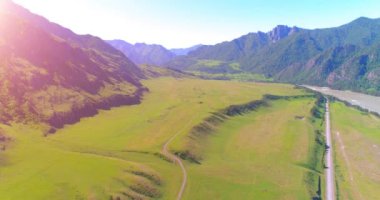 Hava UHD 4K: güneşli yaz sabahında kırsal dağ yolu ve çayır üzerinde havadan uçuş. Ufukta yeşil ağaçlar ve güneş ışınları var. Özgür kırsal asfalt karayolu ve nehir.