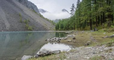 4K UHD dağ gölü zaman ayarlı yaz veya sonbahar saatlerinde. Vahşi doğa ve kırsal dağ vadisi. Çam ağaçlarından oluşan yeşil orman ve gökyüzünde hızlı bulutlar. Motorlu doli kaydırma hareketi