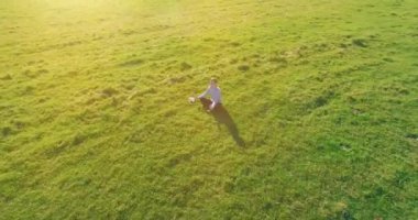Havadan bakış açısı. Yeşil çimlerde oturan ve kırsal alanda not defteri olan bir adamın etrafında alçak yörünge uçuşları. Günlük giysi ve zen meditasyon pozu. Güneş ışınları. Radikal hareket.