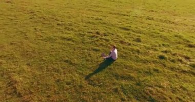 Havadan bakış açısı. Yeşil çimlerde oturan ve kırsal alanda not defteri olan bir adamın etrafında alçak yörünge uçuşları. Günlük giysi ve zen meditasyon pozu. Güneş ışınları. Radikal hareket.