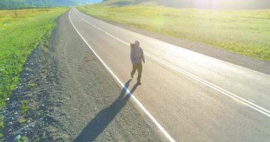 Hava 4K UHD görüntüsü. Asfalt yolda yürüyen otostopçu turist üzerinde alçak irtifa uçuşu. Büyük bir kırsal vadi ve yaz günü güneşli bir çayır. Sırt çantalı gezgin..