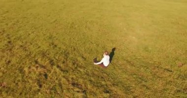 Havadan bakış açısı. Yeşil çimlerde oturan ve kırsal alanda not defteri olan bir adamın etrafında alçak yörünge uçuşları. Günlük giysi ve zen meditasyon pozu. Güneş ışınları. Radikal hareket.