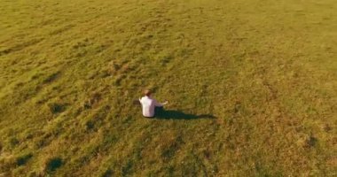 Havadan bakış açısı. Yeşil çimlerde oturan ve kırsal alanda not defteri olan bir adamın etrafında alçak yörünge uçuşları. Günlük giysi ve zen meditasyon pozu. Güneş ışınları. Radikal hareket.
