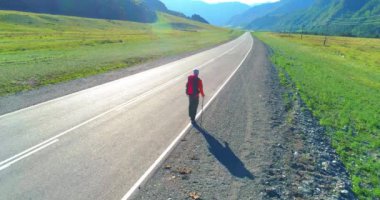 Hava 4K UHD görüntüsü. Asfalt yolda yürüyen otostopçu turist üzerinde alçak irtifa uçuşu. Büyük bir kırsal vadi ve yaz günü güneşli bir çayır. Sırt çantalı gezgin..