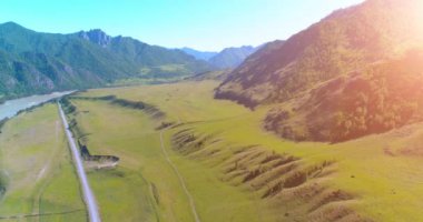 Hava UHD 4K: güneşli yaz sabahında kırsal dağ yolu ve çayır üzerinde havadan uçuş. Ufukta yeşil ağaçlar ve güneş ışınları var. Özgür kırsal asfalt karayolu ve nehir.