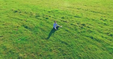 Havadan bakış açısı. Yeşil çimlerde oturan ve kırsal alanda not defteri olan bir adamın etrafında alçak yörünge uçuşları. Günlük giysi ve zen meditasyon pozu. Güneş ışınları. Radikal hareket.