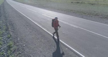 Hava 4K UHD görüntüsü. Asfalt yolda yürüyen otostopçu turist üzerinde alçak irtifa uçuşu. Büyük bir kırsal vadi ve yaz günü güneşli bir çayır. Çiğ düz renk, düzeltme yok.