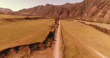 Hava UHD 4K: güneşli yaz sabahında kırsal dağ yolu ve çayır üzerinde havadan uçuş. Ufukta yeşil ağaçlar ve güneş ışınları var. Özgür kırsal asfalt karayolu ve nehir.