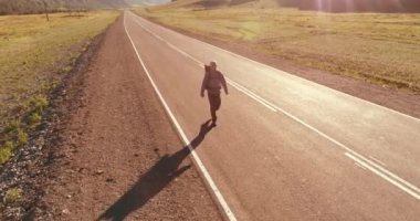 Hava 4K UHD görüntüsü. Asfalt yolda yürüyen otostopçu turist üzerinde alçak irtifa uçuşu. Büyük bir kırsal vadi ve yaz günü güneşli bir çayır. Sırt çantalı gezgin..