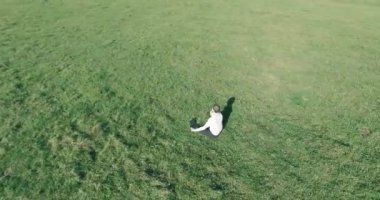 Havadan bakış açısı. Yeşil çimlerde oturan ve kırsal alanda not defteri olan bir adamın etrafında alçak yörünge uçuşları. Günlük giysi ve zen meditasyon pozu. Güneş ışınları. Radikal hareket.