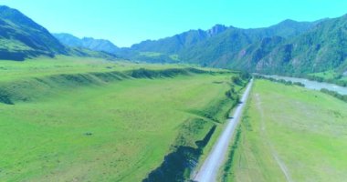 Hava UHD 4K: güneşli yaz sabahında kırsal dağ yolu ve çayır üzerinde havadan uçuş. Ufukta yeşil ağaçlar ve güneş ışınları var. Özgür kırsal asfalt karayolu ve nehir.