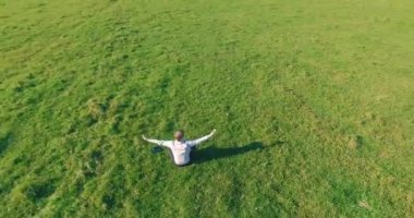 Havadan bakış açısı. Yeşil çimlerde oturan ve kırsal alanda not defteri olan bir adamın etrafında alçak yörünge uçuşları. Gündelik kıyafet ve Zen meditasyonu. Eller havada. Güneş ışınları. Radikal hareket.