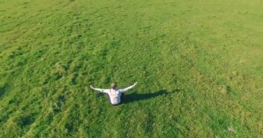 Havadan bakış açısı. Yeşil çimlerde oturan ve kırsal alanda not defteri olan bir adamın etrafında alçak yörünge uçuşları. Gündelik kıyafet ve Zen meditasyonu. Eller havada. Güneş ışınları. Radikal hareket.
