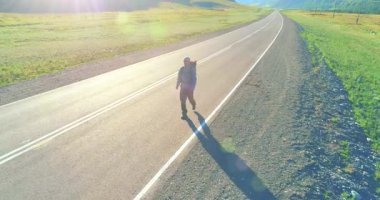 Hava 4K UHD görüntüsü. Asfalt yolda yürüyen otostopçu turist üzerinde alçak irtifa uçuşu. Büyük bir kırsal vadi ve yaz günü güneşli bir çayır. Sırt çantalı gezgin..