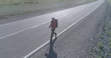 Hava 4K UHD görüntüsü. Asfalt yolda yürüyen otostopçu turist üzerinde alçak irtifa uçuşu. Büyük bir kırsal vadi ve yaz günü güneşli bir çayır. Çiğ düz renk, düzeltme yok.