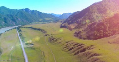 Hava UHD 4K: güneşli yaz sabahında kırsal dağ yolu ve çayır üzerinde havadan uçuş. Ufukta yeşil ağaçlar ve güneş ışınları var. Özgür kırsal asfalt karayolu ve nehir.