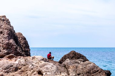 Adam kayanın üzerinde oturuyor ve denize bakıyor.