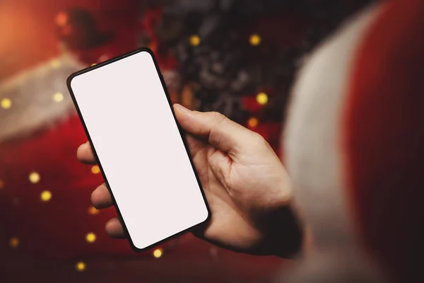 戴着圣诞礼帽的男人手里拿着手机 屏幕空白 背景是圣诞装饰品 网上购物和做广告 — 图库照片