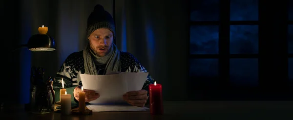 stock image shocked man in winter clothes looks at household expenses bills while sitting in candlelight in cold room. energy crisis, heating season