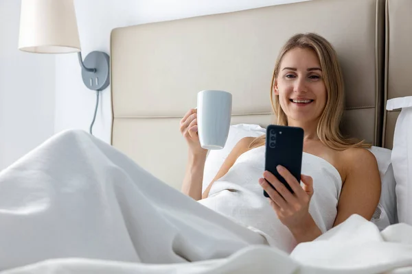 Sorrindo Jovem Deitado Cama Usando Telefone Enquanto Bebe Café Manhã — Fotografia de Stock