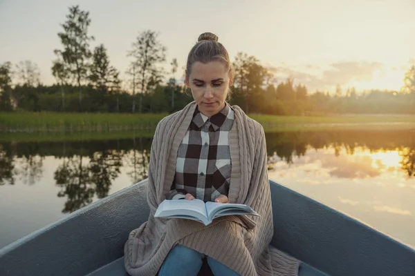 湖の上のボートに座って日没時に本を読む肩に毛布を持つ女性 — ストック写真