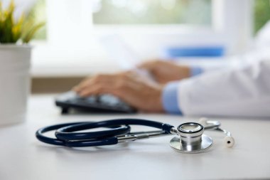Klinikteki masada steteskop var. Doktor arka planda bilgisayar üzerinde çalışıyor