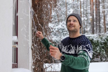 Evin dış dekorasyonunu yapan adam, karlı kış gününde Noel ışıklarıyla süslüyor.
