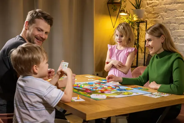 有两个孩子的家庭在一起消磨时光 在家里坐在桌旁玩棋盘游戏 联合活动 免版税图库图片