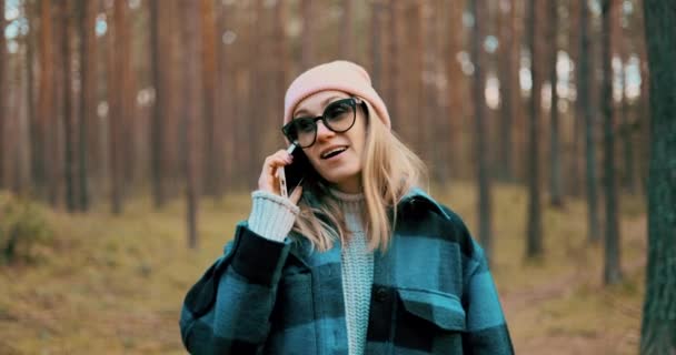 寒冷的日子里 一位迷人的年轻女子一边在松树林中散步一边用手机聊天 — 图库视频影像