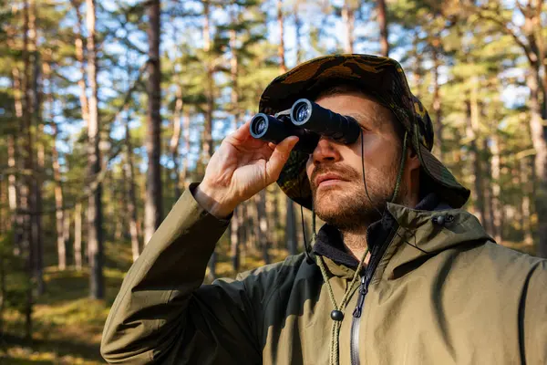 身着迷彩服的男人透过双筒望远镜在森林里寻找 停车场监测 图库图片