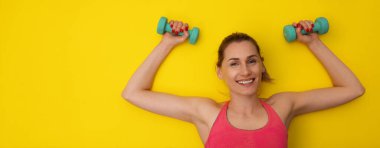 Gülümseyen fitness kadını, sarı jimnastik paspasının üzerine uzanmış, ellerinde halterlerle. sağlıklı aktif yaşam tarzı. Kopyalama alanı olan pankart