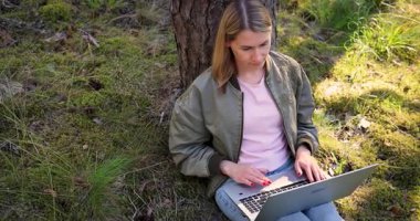 Ormanda dizüstü bilgisayar kullanan serbest çalışan bir kadın. Dijital göçebe, küçük işletme sahibi ağaca karşı oturuyor ve doğada uzaktan çalışıyor. Serbest kumanda çalışma konsepti