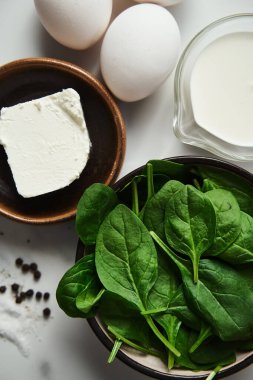 Sağlıklı ıspanak ve feta omleti için malzemeler. Ağır kremalı yumurtalar, siyah seramik kasede taze ıspanak yaprakları, beyaz mermer arka plan. Yüksek kalite fotoğraf