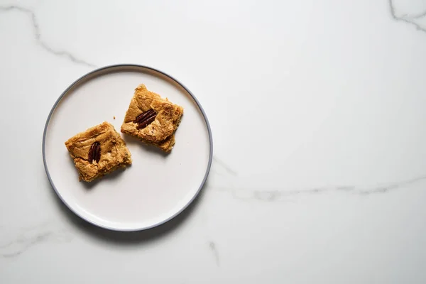 Two squares of Blondie cookies with pecan nuts on white plate. White chocolate sweet and tasty, chewy cookie bars. Top view, copy space. High quality photo