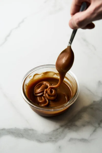 Salted Caramel sauce in glass bowl on white marble background. The hand holding the spoon dips into the caramel sauce, the caramel stretches. Butter, sugar with cream and salt. High quality photo