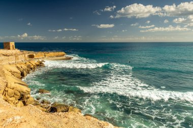 Canlı turkuaz bir denizi gören altın kumtaşı katmanlarıyla muhteşem bir sahil kayalığı manzarası. Güneş ışığı kayaların engebeli dokusunu vurgular..
