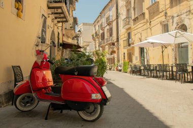 A bright red vintage scooter parked on a charming narrow street in a Sicilian town, surrounded by historic buildings, outdoor caf seating.