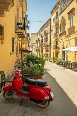 A bright red vintage scooter parked on a charming narrow street in a Sicilian town, surrounded by historic buildings, outdoor caf seating.