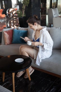 Lobide oturan kız kahve içer ve telefona bakar.