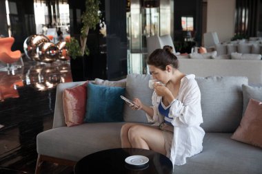 Lobide oturan kız kahve içer ve telefona bakar.