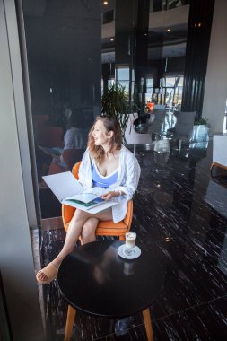 Bir kız lobide oturuyor, kahve içiyor ve bir dergiye bakıyor.