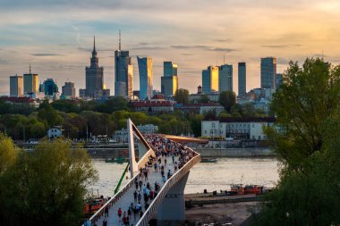 Warszawa, Polonya - 9 Nisan 2024: Varşova şehir merkezinin panoramik manzarası, yeni açılan yaya ve bisiklet köprüsünün modern şekli tarafından çerçevelenmiş, canlı bir anıt ve modern gökdelenler karışımına sahip..