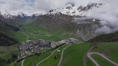 Andermatt 'ın yeşil çayırlarla çevrili dağlık köyü ve arka planda karlı tepeler, Uri Kantonu, İsviçre, Avrupa