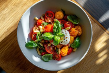 Kiraz domatesli renkli bir salata, mozarella topları ve ahşap bir masada beyaz bir kasede fesleğen yaprakları..